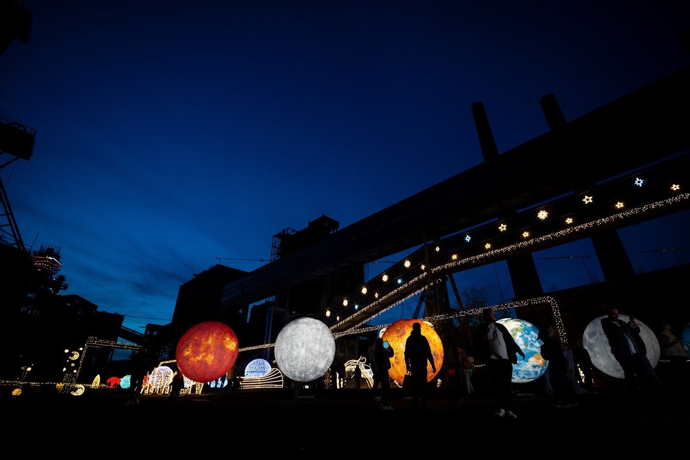 Noční expozice svítících planet s procházejícími návštěvníky v industriálním areálu.